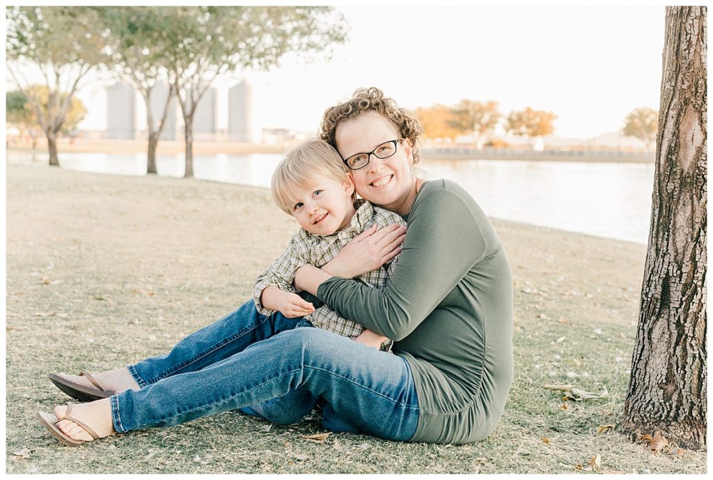 Mom hugging little boy while sitting next to a tree and a lake at Morrison Ranch