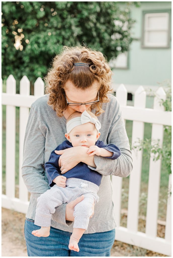 mom kissing her new born baby girls head in front of a white picket fence at Tumbleweed Park 