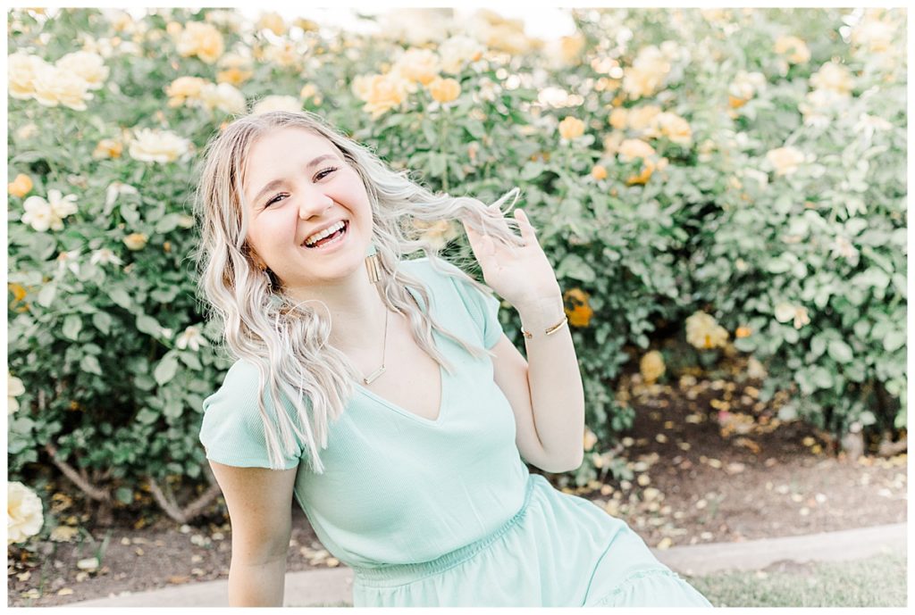 girl sitting in front of yellow rose bushes laughing and playing with her hair